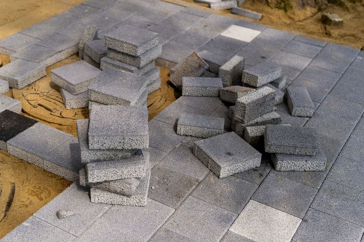 A stack of square grey pavers sits on a sand base, surrounded by partially laid paving stones on a construction site.