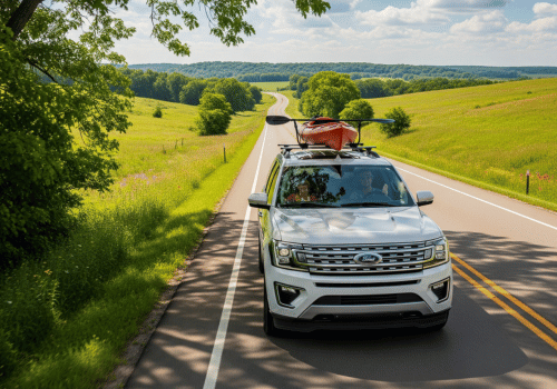 White SUV with rooftop kayaks driving on a winding country road through green hills under a blue sky