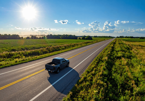Pickup truck driving down a rural highway through open farmland under bright sunshine and blue skies