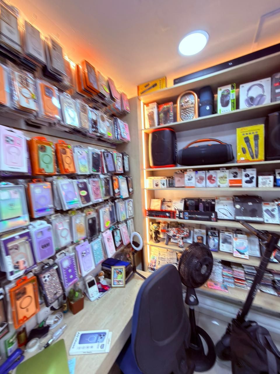 Interior de una pequeña tienda con estantes de accesorios para teléfonos y una persona sentada trabajando en un escritorio.