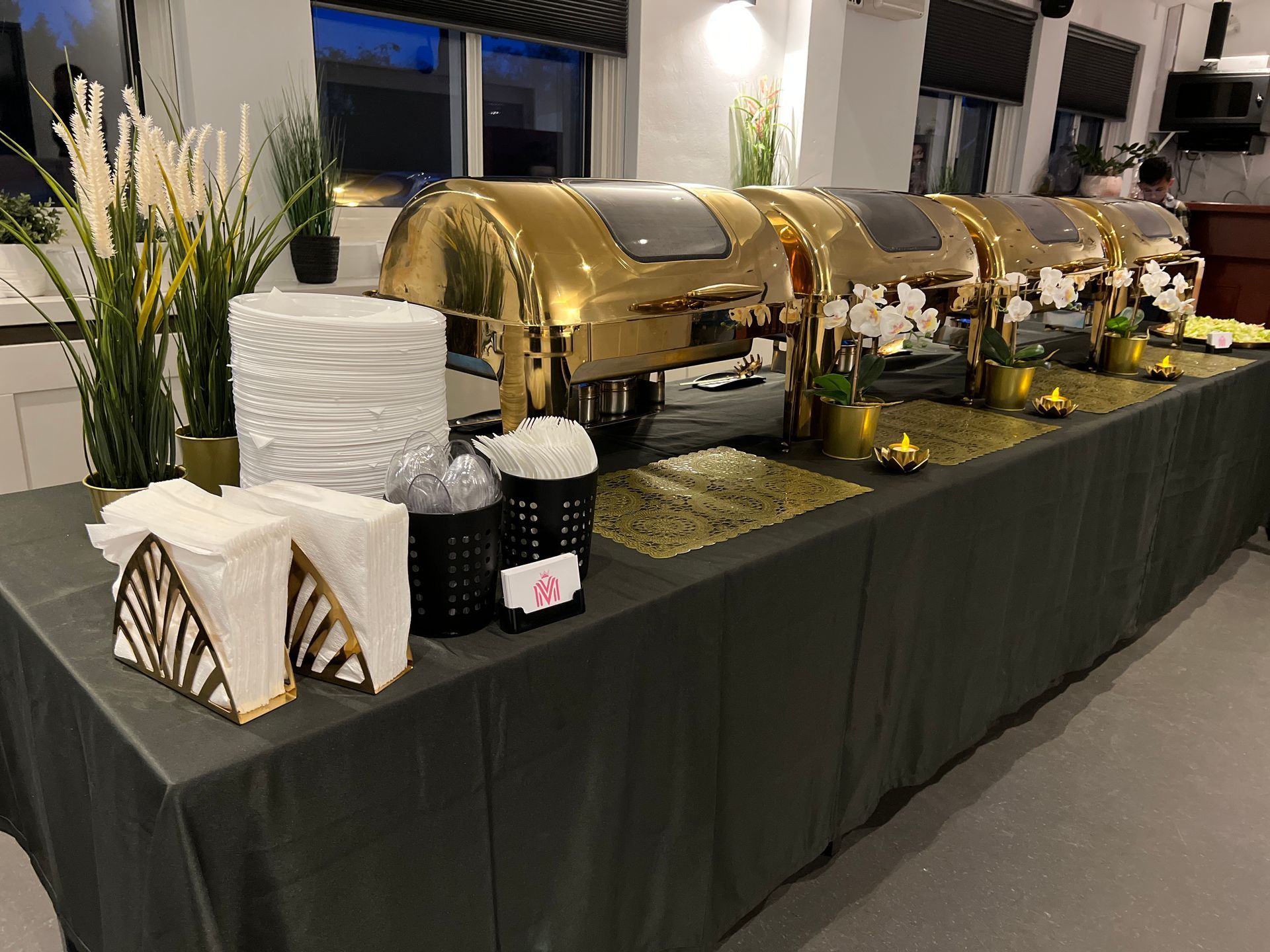Buffettafel met gouden warmhoudschalen, witte borden, servetten en bloemversieringen.