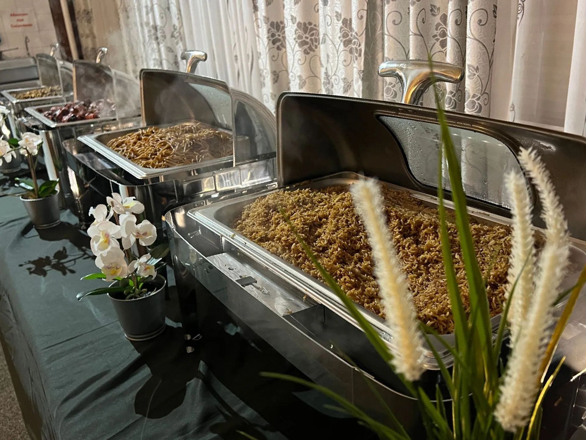Buffettafel met diverse zilveren warmhoudschalen gevuld met eten, versierd met witte bloemen en grassen.