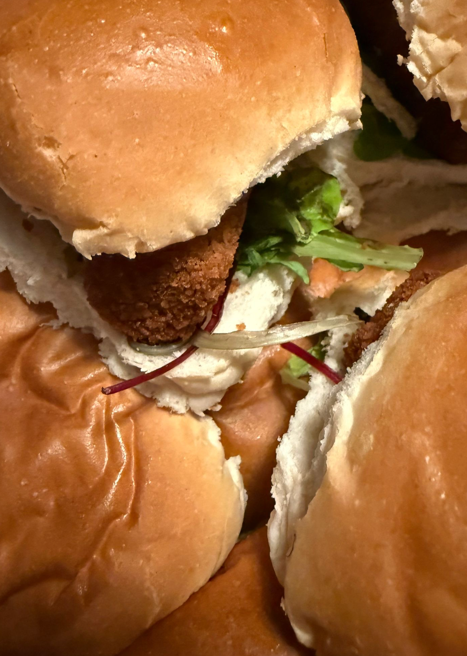 Close-up van een aantal kleine sliders op witte broodjes met een gebakken burger, sla en een paar rode sliertjes.
