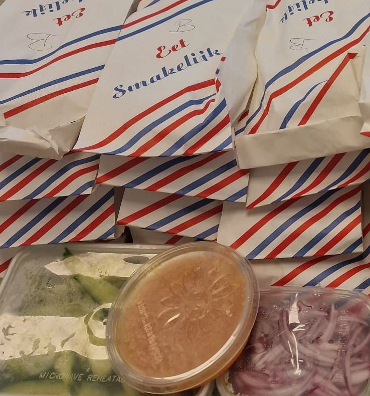 Tassen met rode, witte en blauwe strepen, volgestapeld met bakjes eten, waaronder salade, saus en uien.