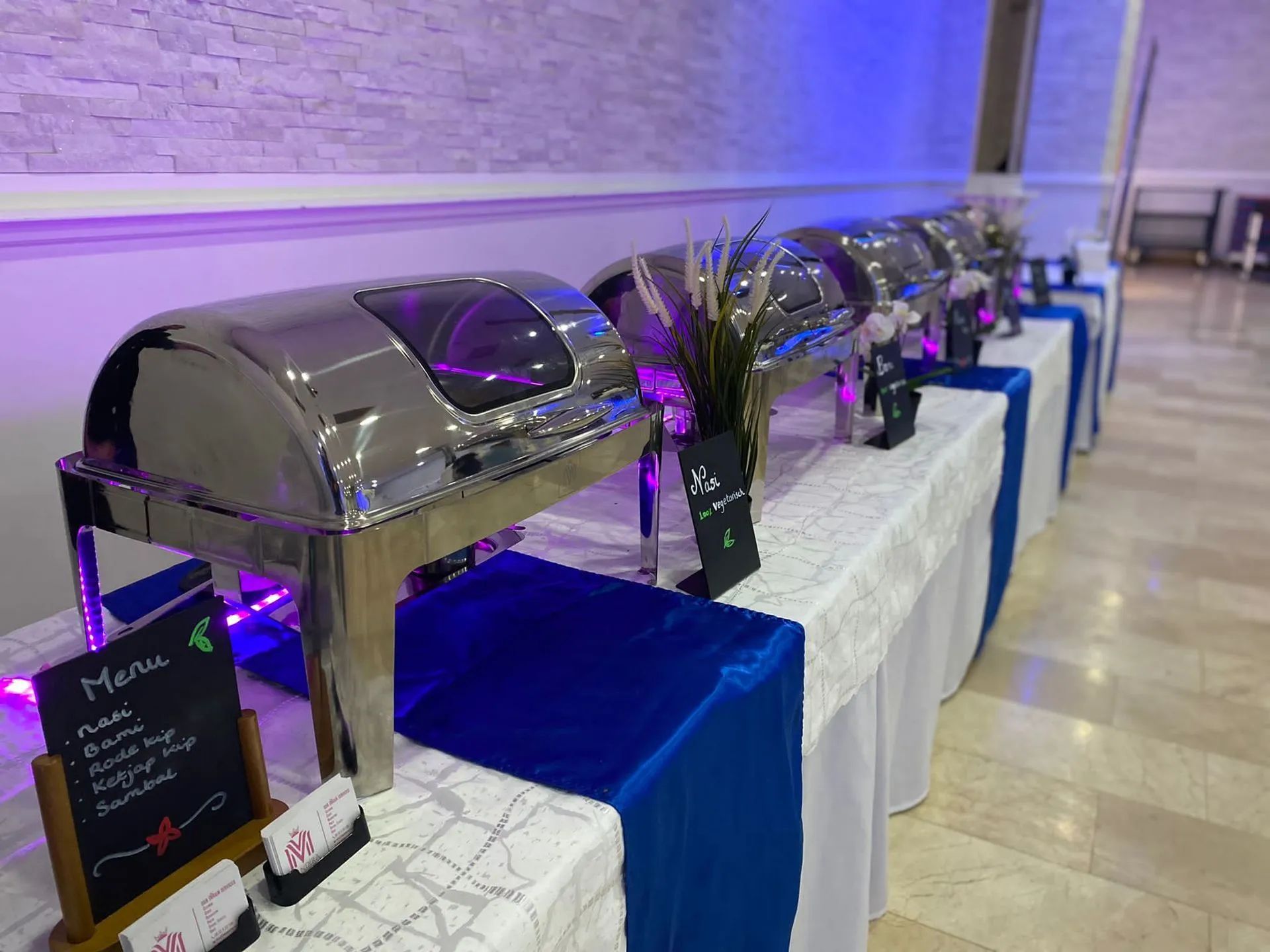 Buffet met zilverkleurige warmhoudschalen op witte en blauwe tafelkleden; menu op een krijtbord.
