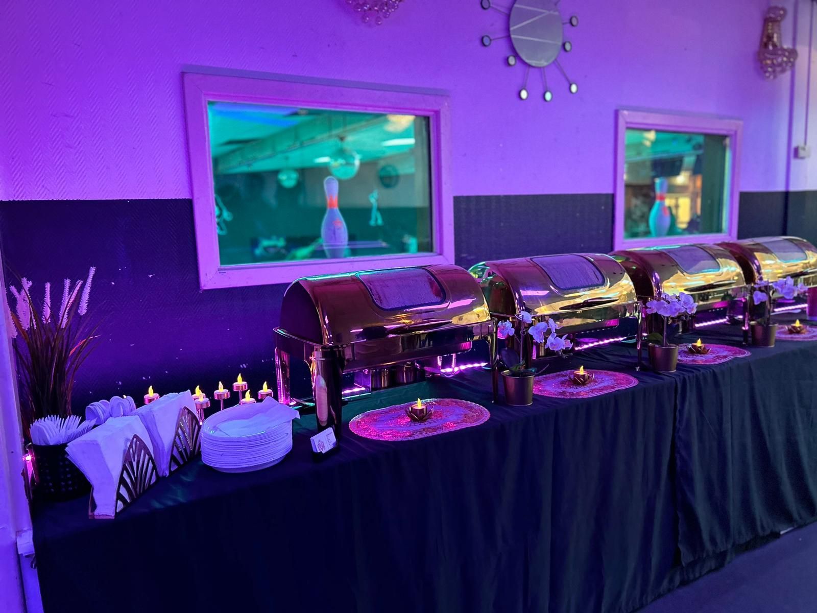 Buffettafel met gouden warmhoudschalen, paarse verlichting en versieringen in een evenementenlocatie.