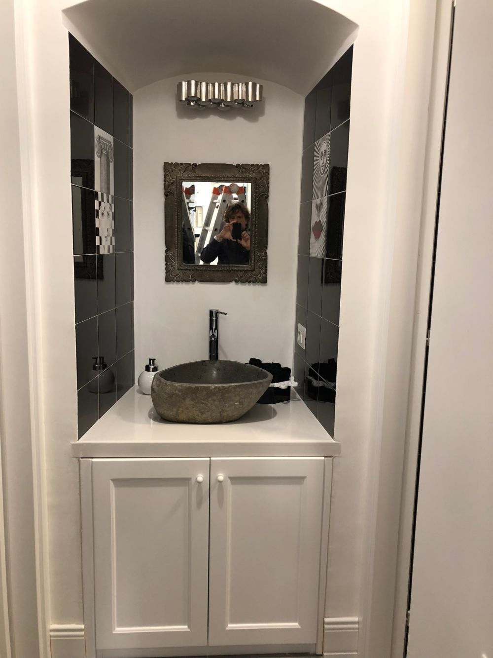 Compact bathroom vanity nook with white cabinets, stone sink, mirror, and dark tile walls