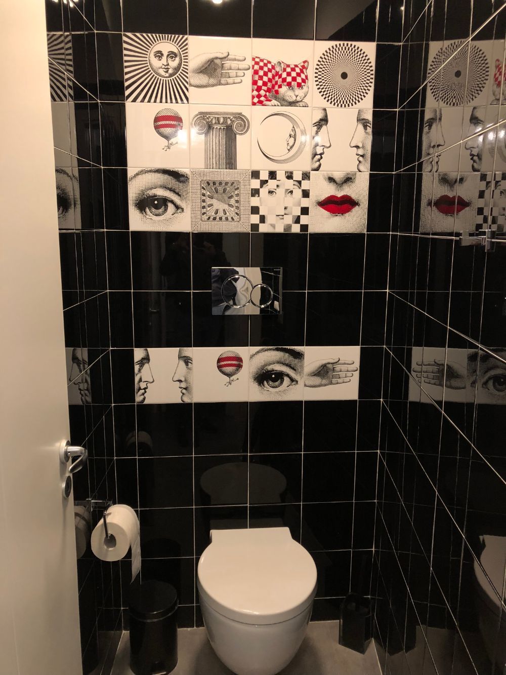 Small toilet room with black tile walls, a white toilet, and black-and-white pop-art tiles above it.