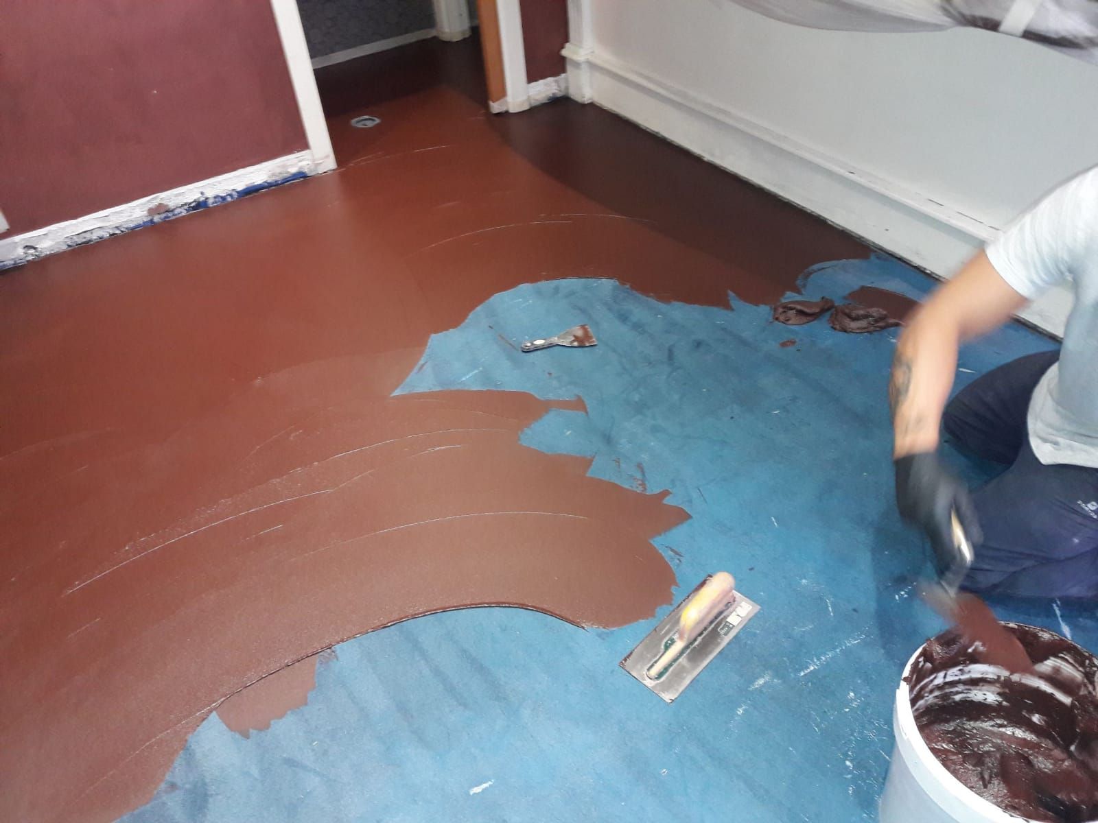 Person painting a floor blue in a room, with a paint bucket and roller nearby.