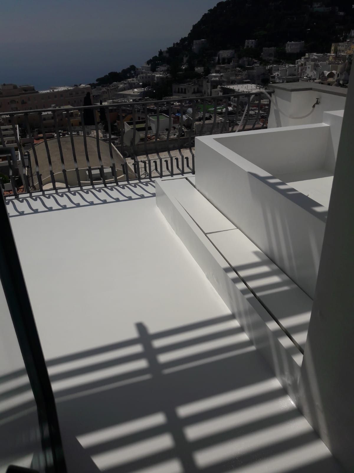 White rooftop terrace with metal railing, sunlit stairs, and a coastal hillside view in the distance