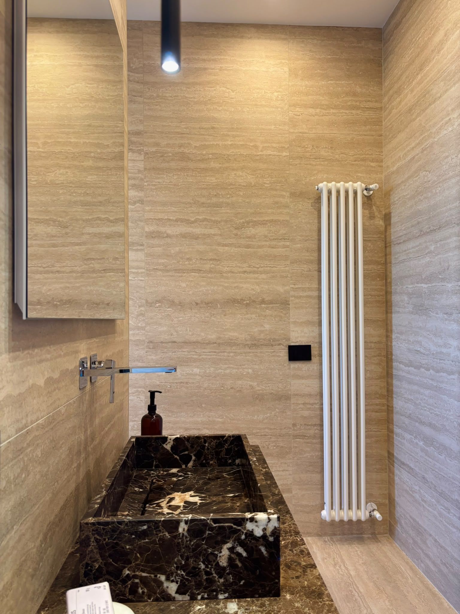 Modern bathroom with beige tile walls, black sink vanity, round mirror, and white radiator under a hanging light