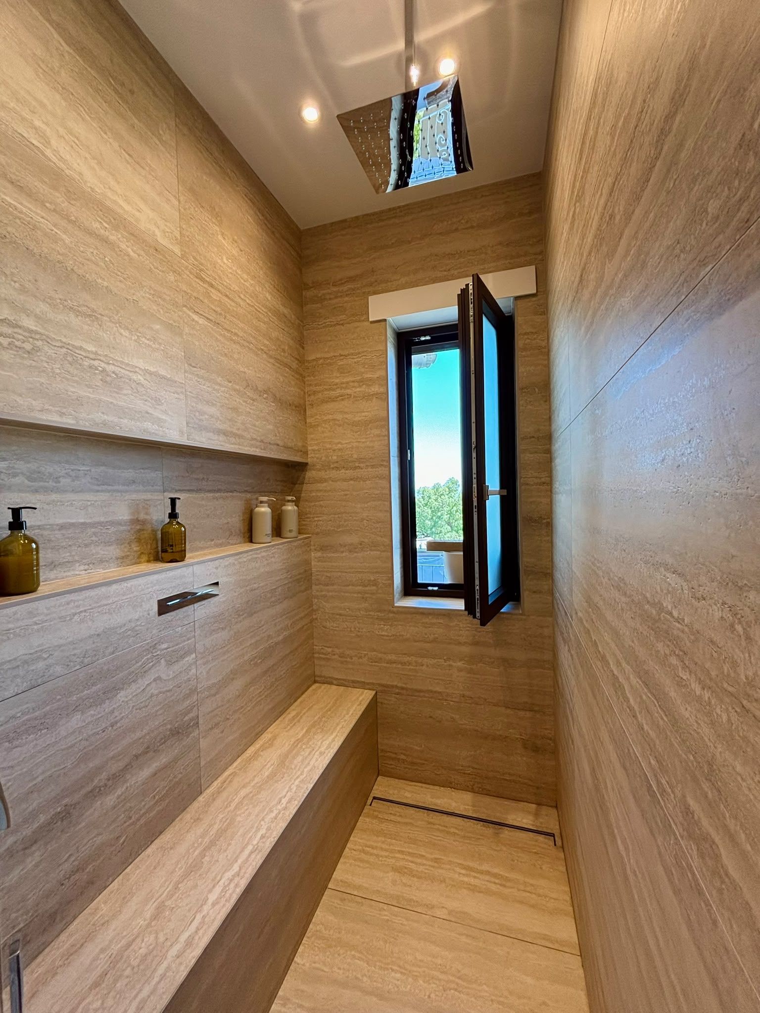 Narrow beige tiled shower with built-in bench, wall niches, and an open window above the doorway.