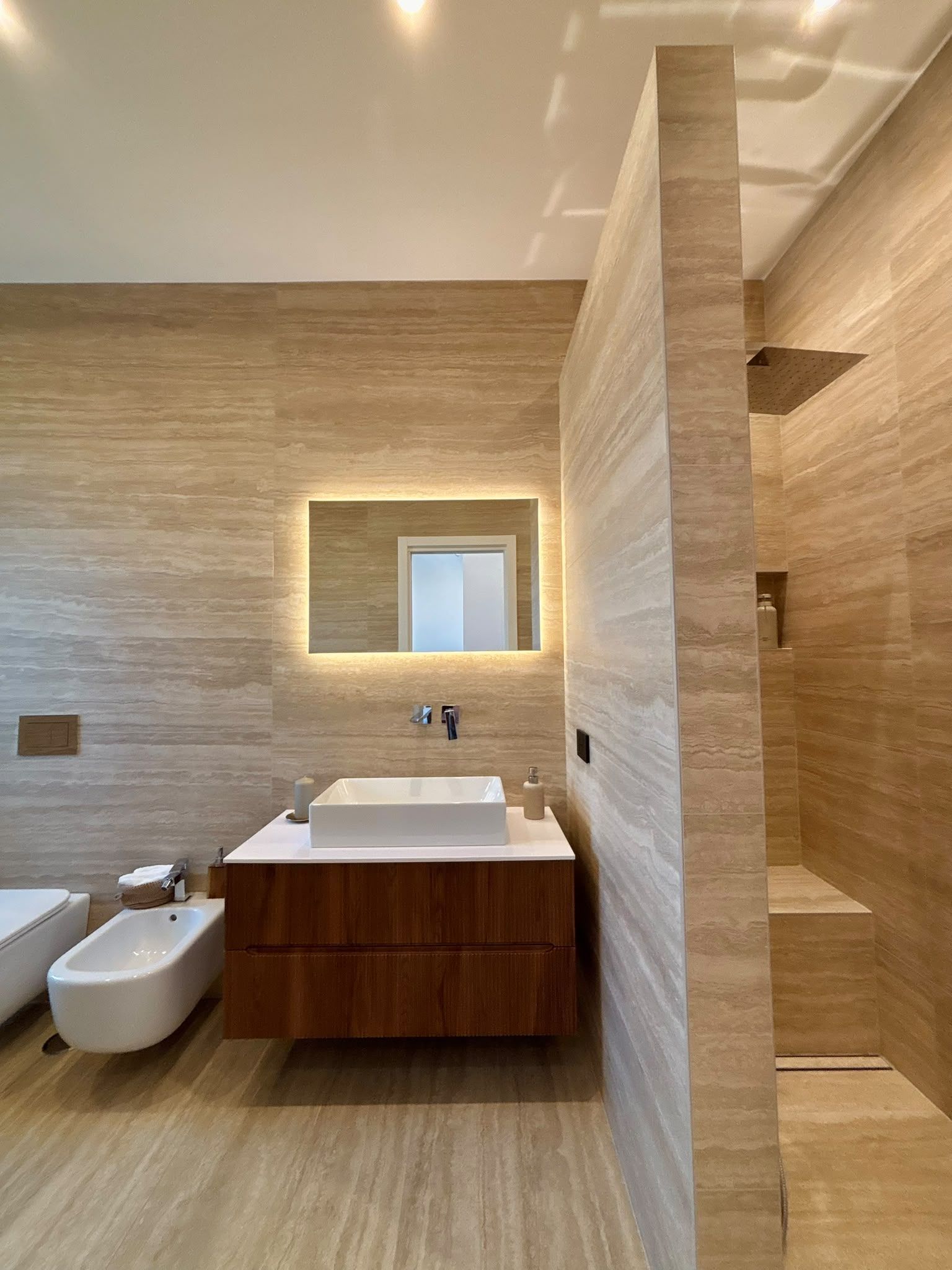 Modern bathroom with wood vanity, wall-mounted sink, toilet, and walk-in shower with beige tile.