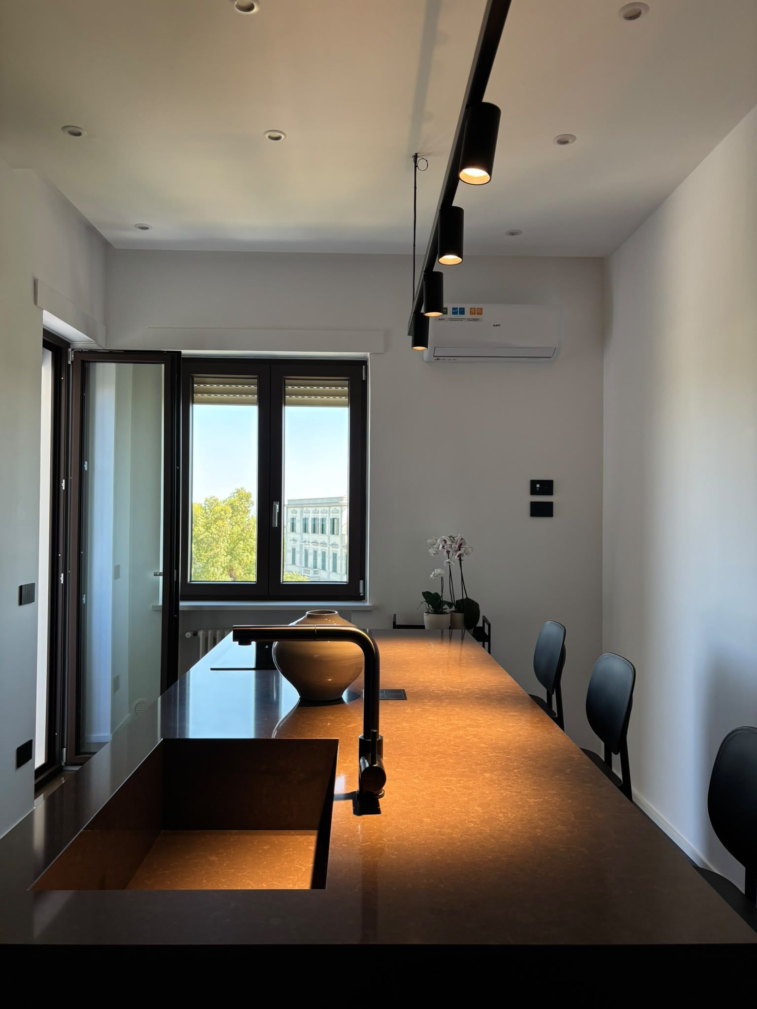 Modern minimalist room with long table, black chairs, track lighting, and a window view of buildings.