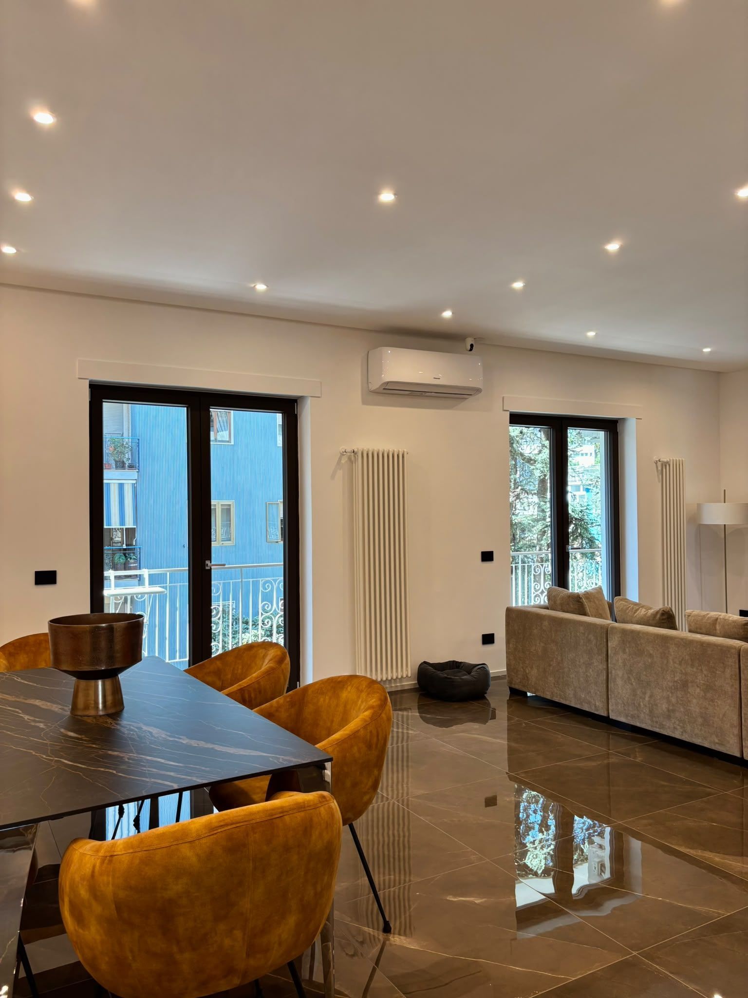 Modern open-plan dining room with black table, mustard chairs, beige sofa, and glass doors.