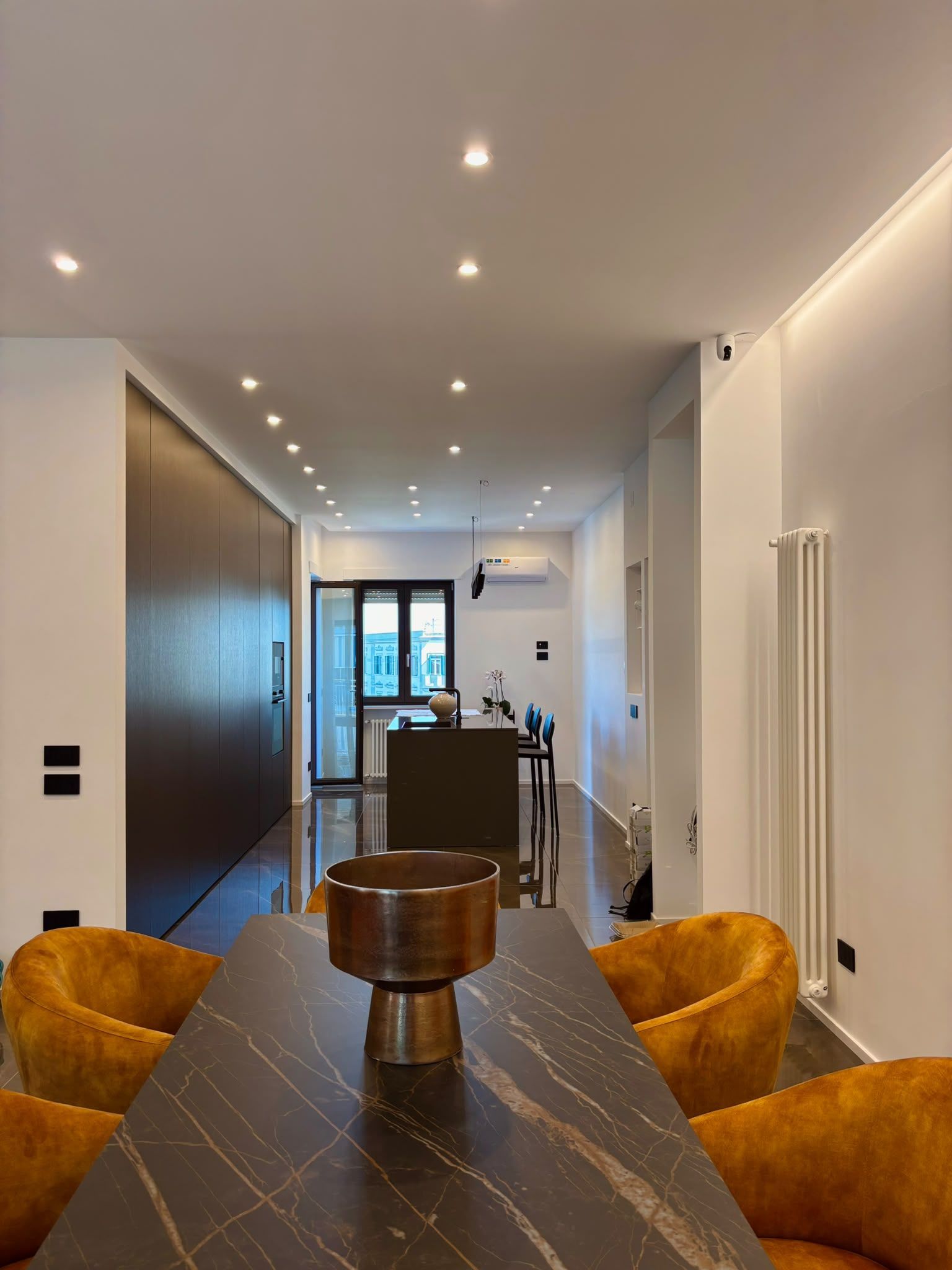 Modern hallway dining area with marble table, mustard chairs, recessed lighting, and large window at the end
