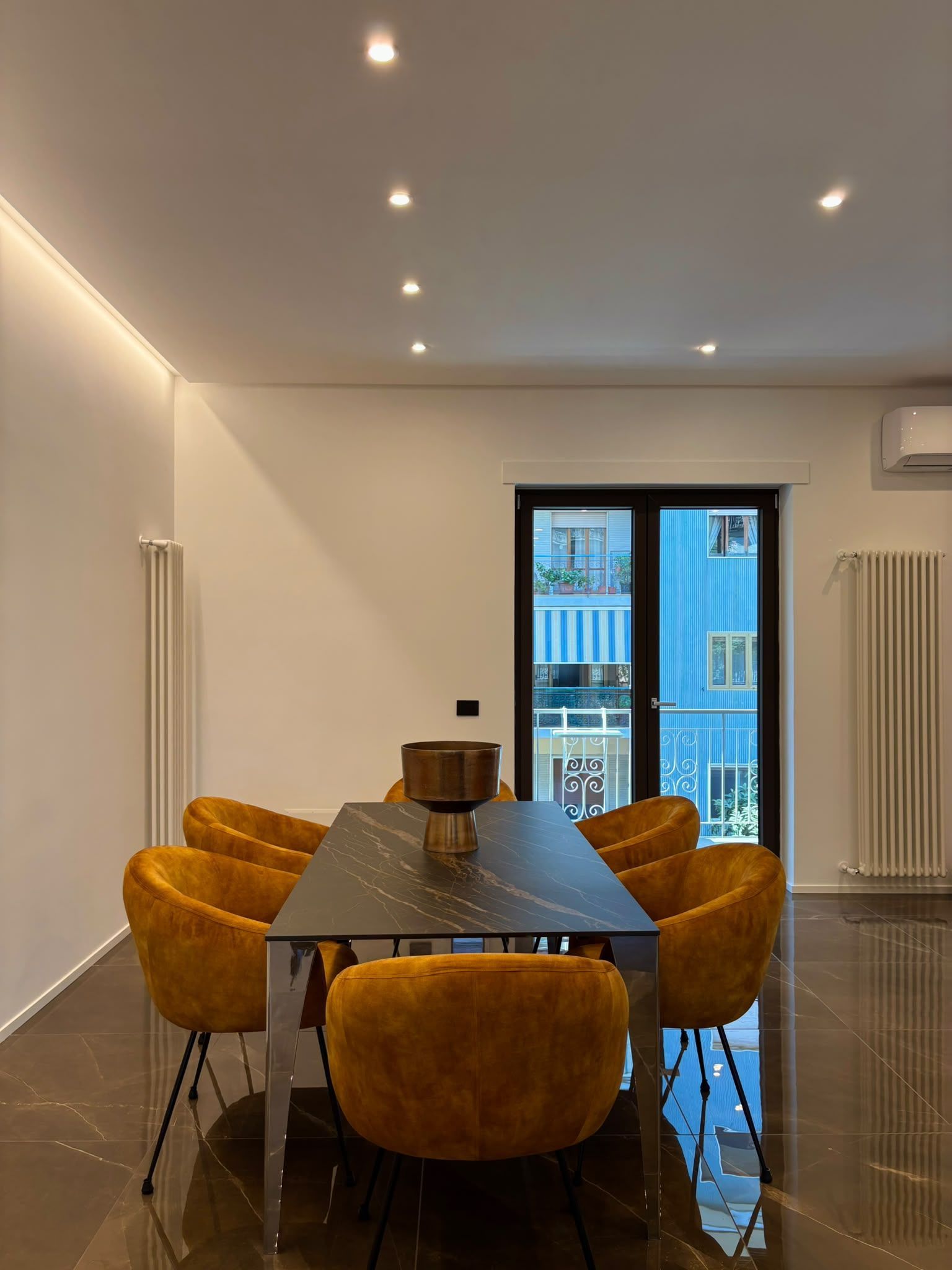 Modern dining room with mustard chairs, long table, white walls, and glass doors to a balcony.