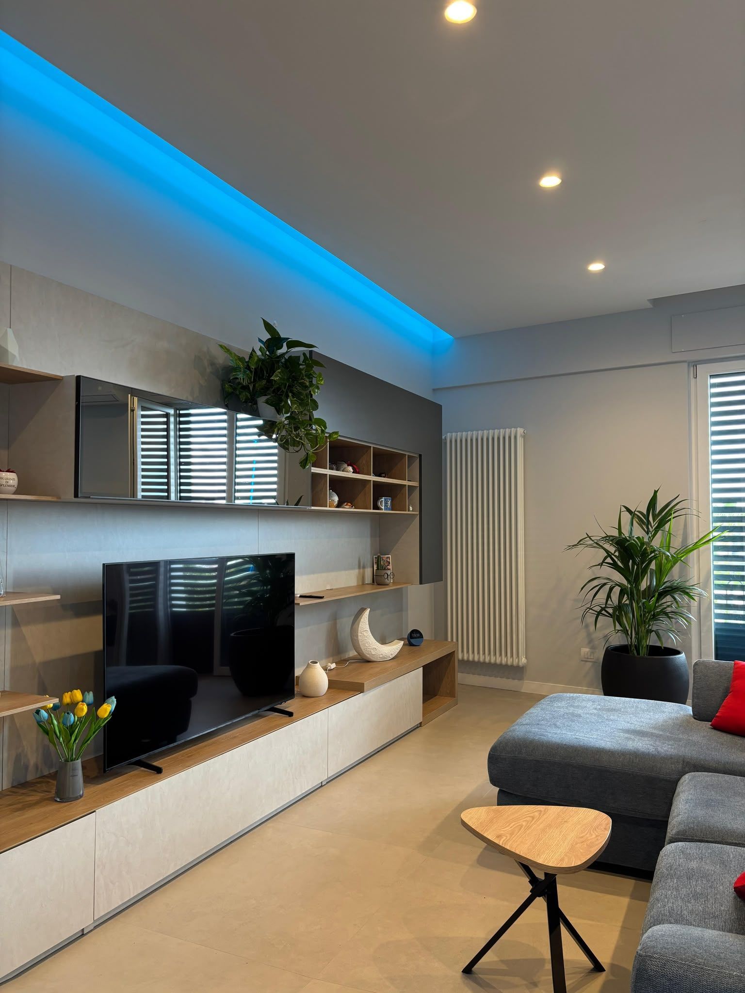 Modern living room with blue LED lighting, TV cabinet, gray sofa, plants, and a skylight window.