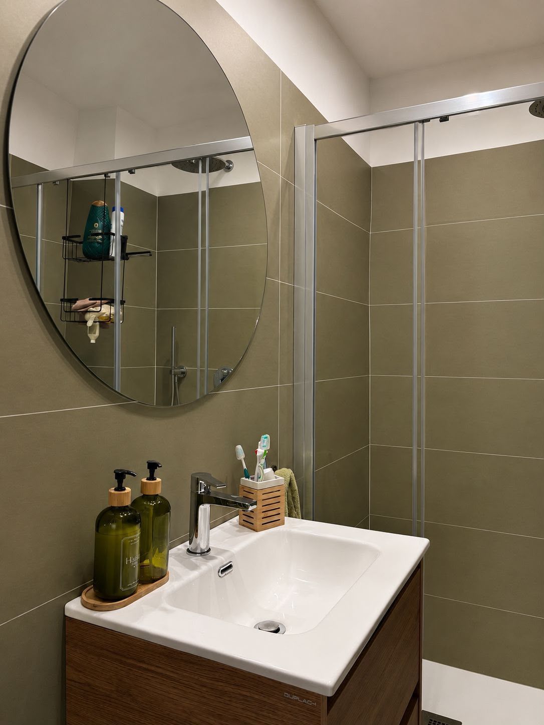 Bathroom sink with oval mirror, white countertop, and glass shower enclosure.