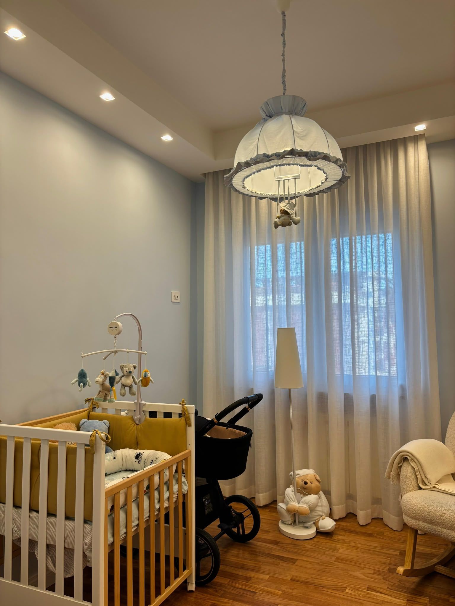 Nursery with crib, stroller, teddy bear, and hanging lamp by a window with sheer curtains.