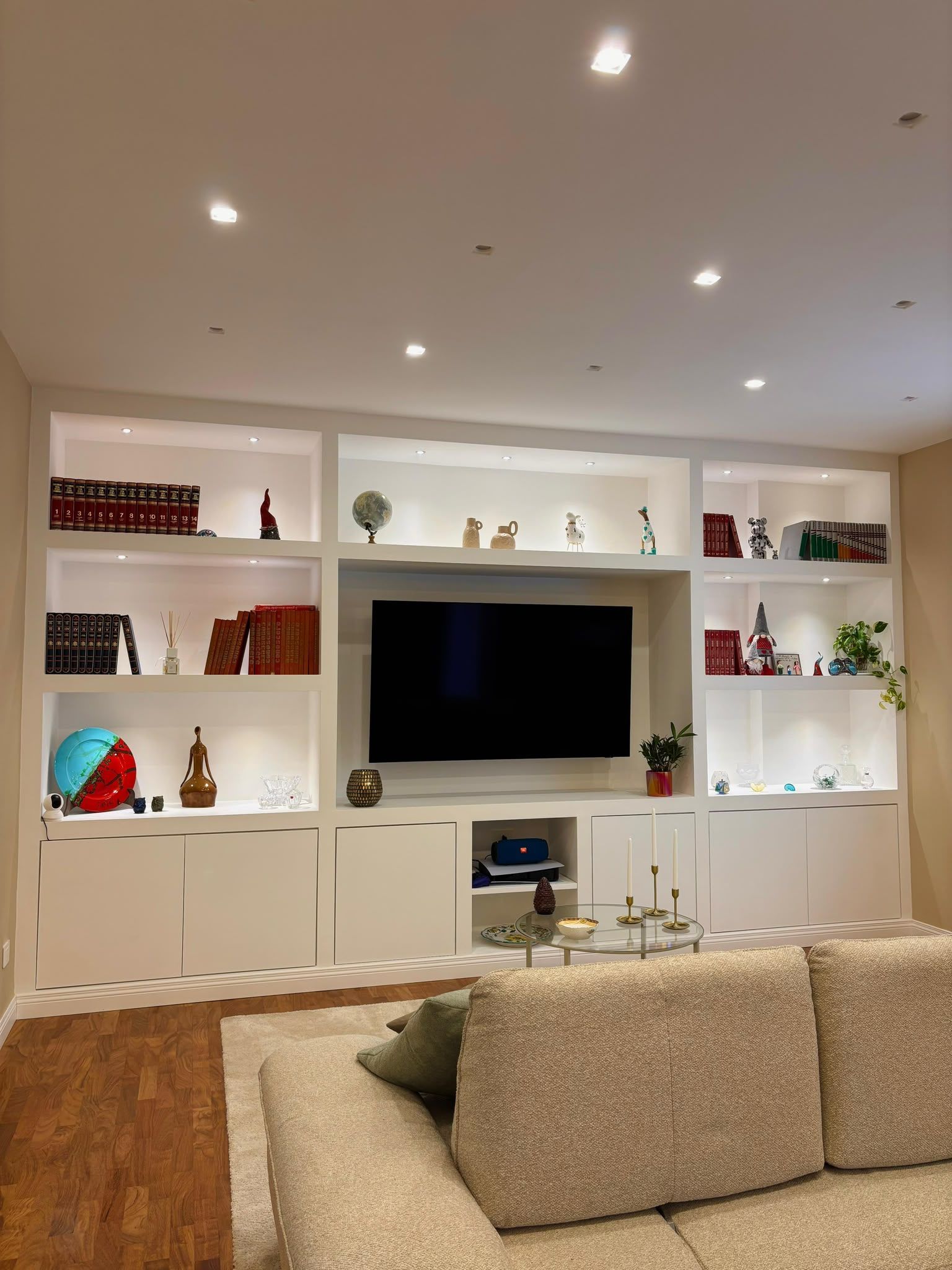 Bright living room with built-in white shelves, a wall-mounted TV, recessed lights, and a beige sofa in front.