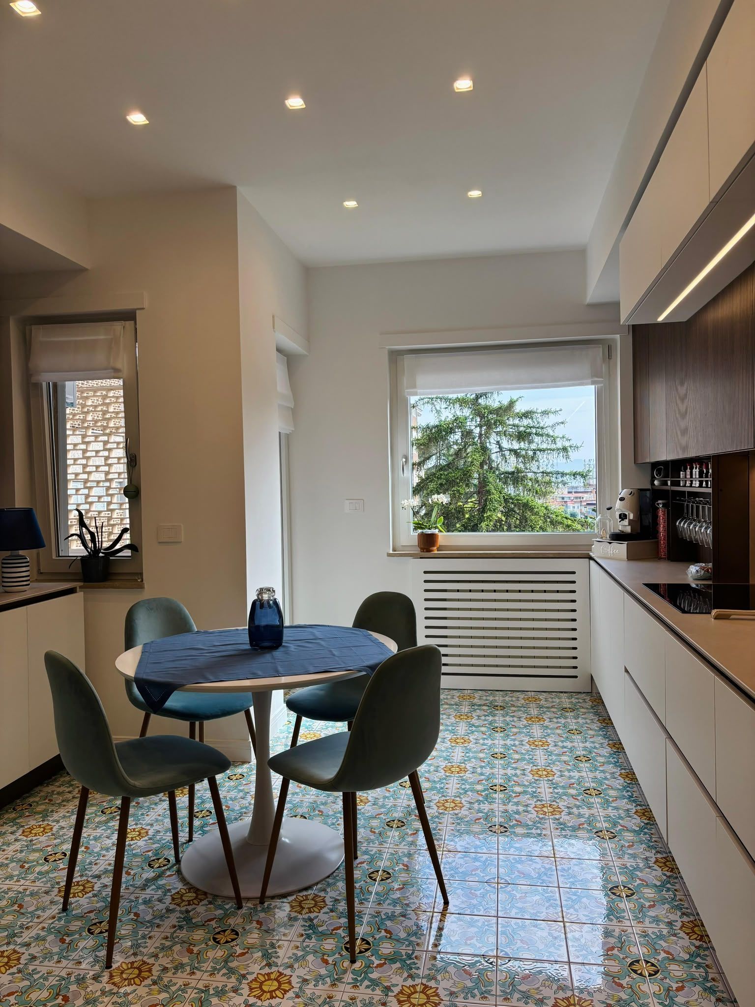 Modern kitchen with round table, dark chairs, patterned tile floor, and bright window with plants