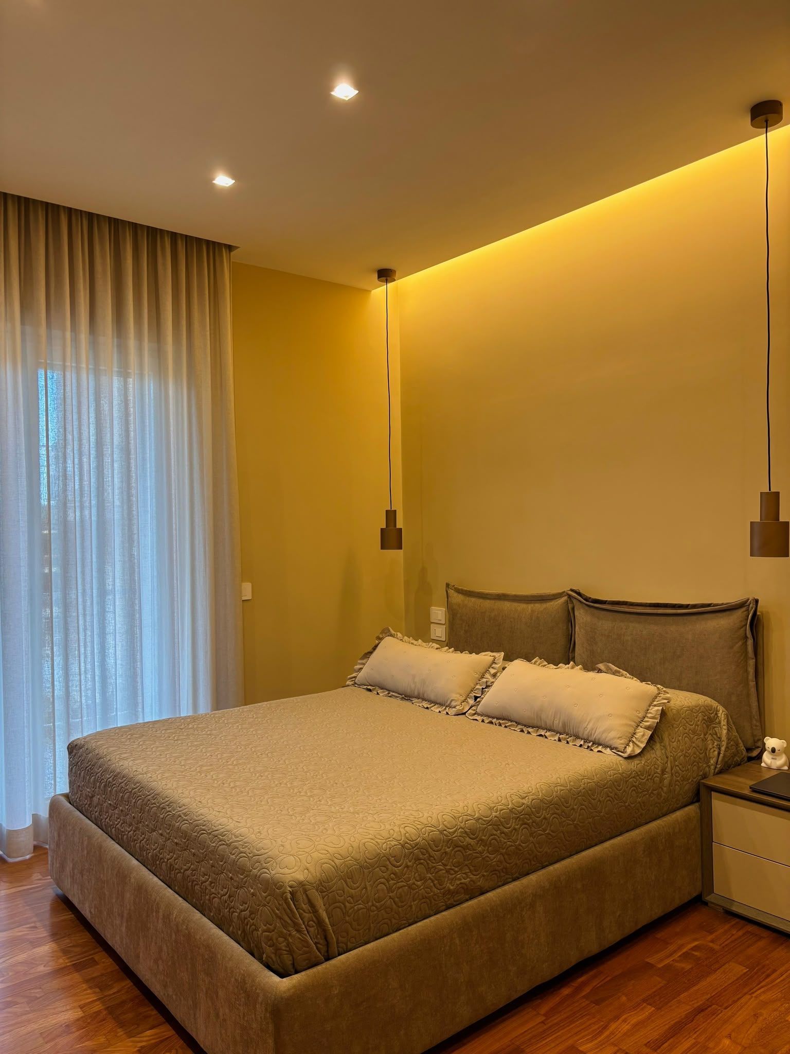 Modern bedroom with a beige bed, warm wall lighting, and sheer curtains by the window