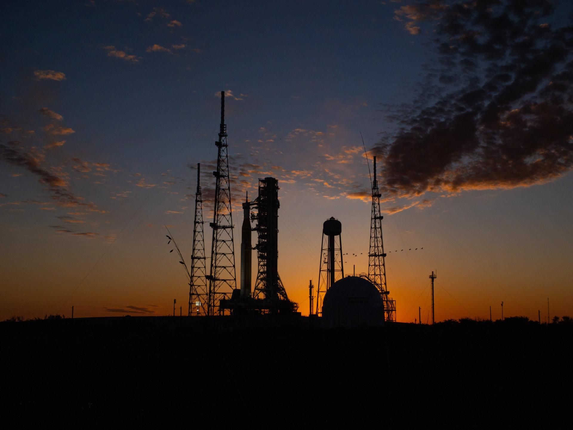 A rocket and spacecraft standing on the launch pad as the sun sets behind them