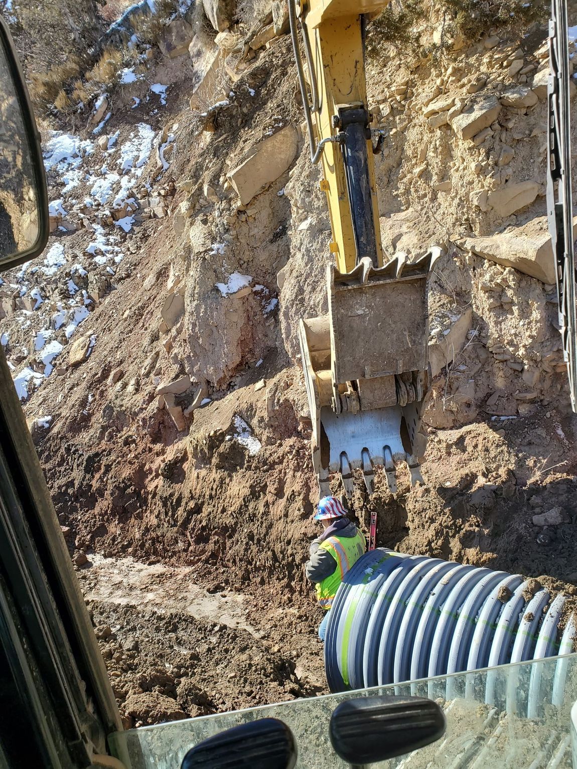 Excavator digging a muddy trench beside a corrugated pipe and steep dirt wall