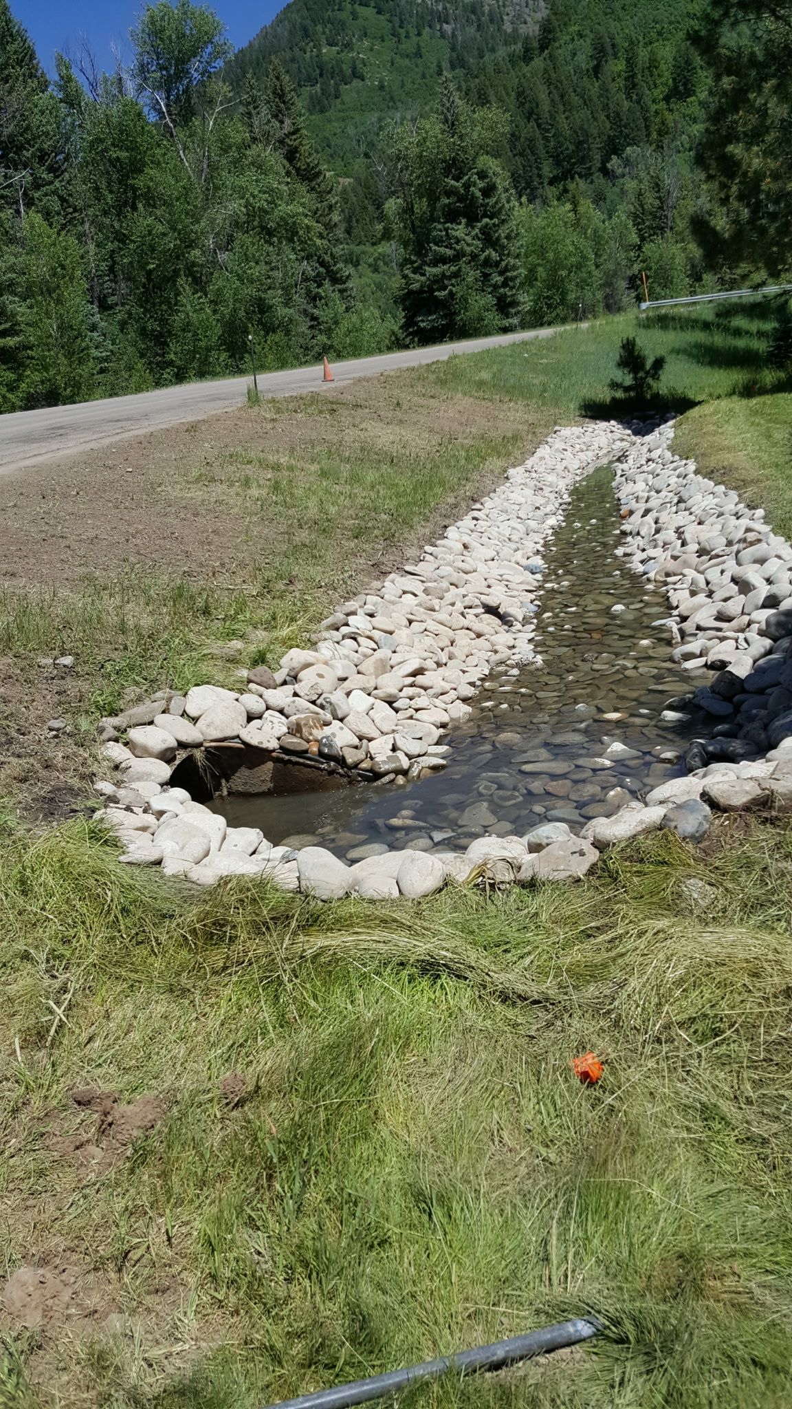 Stone-lined drainage ditch with flowing water beside a grassy roadside and trees