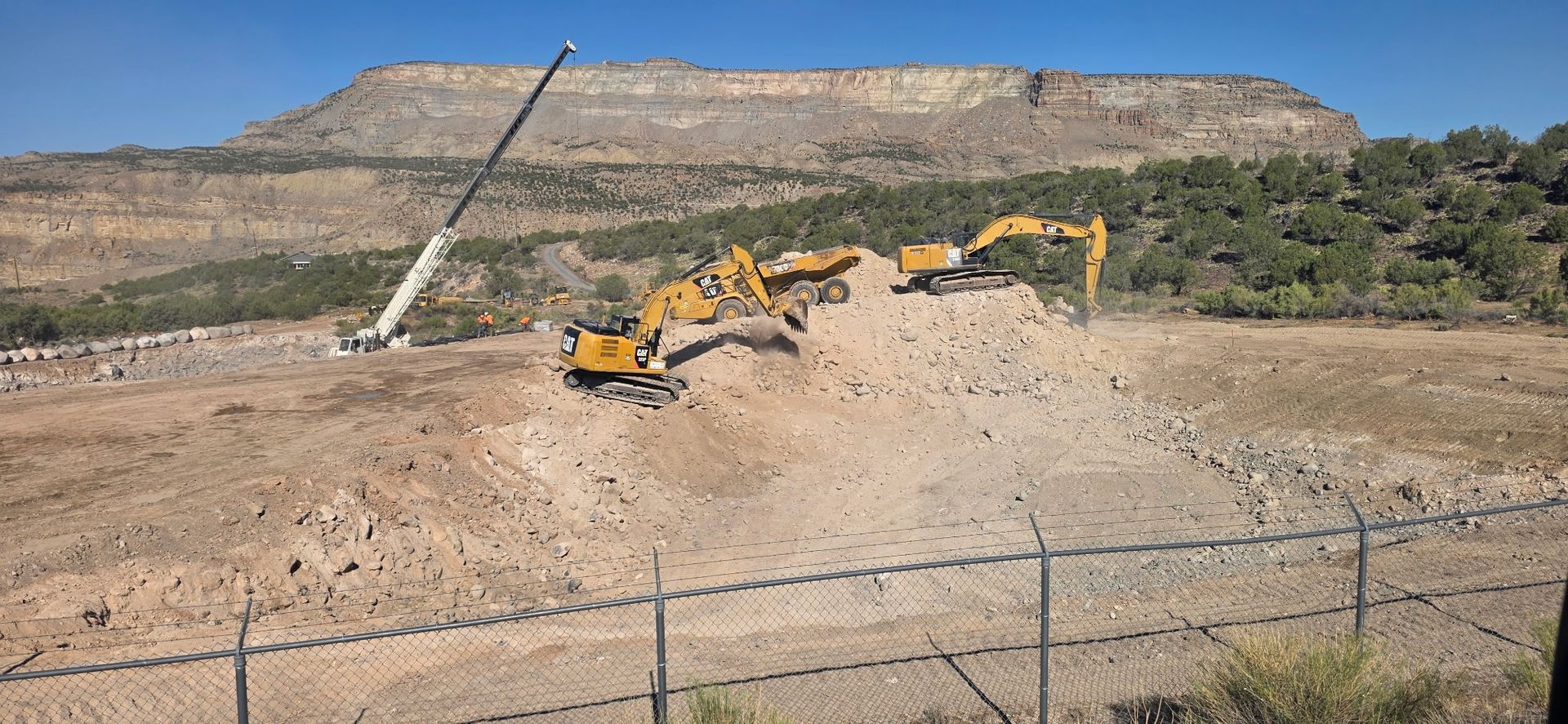 Excavators working on a dirt hillside with a rocky plateau and trees in the background
