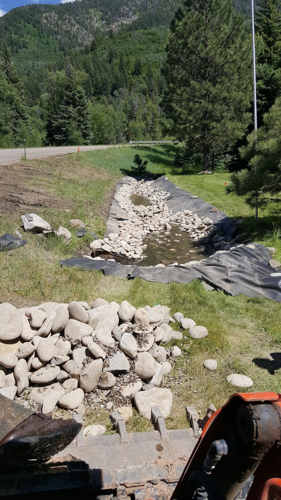 Rocky stream bed with piled stones, grassy banks, and a forested hillside in the background.