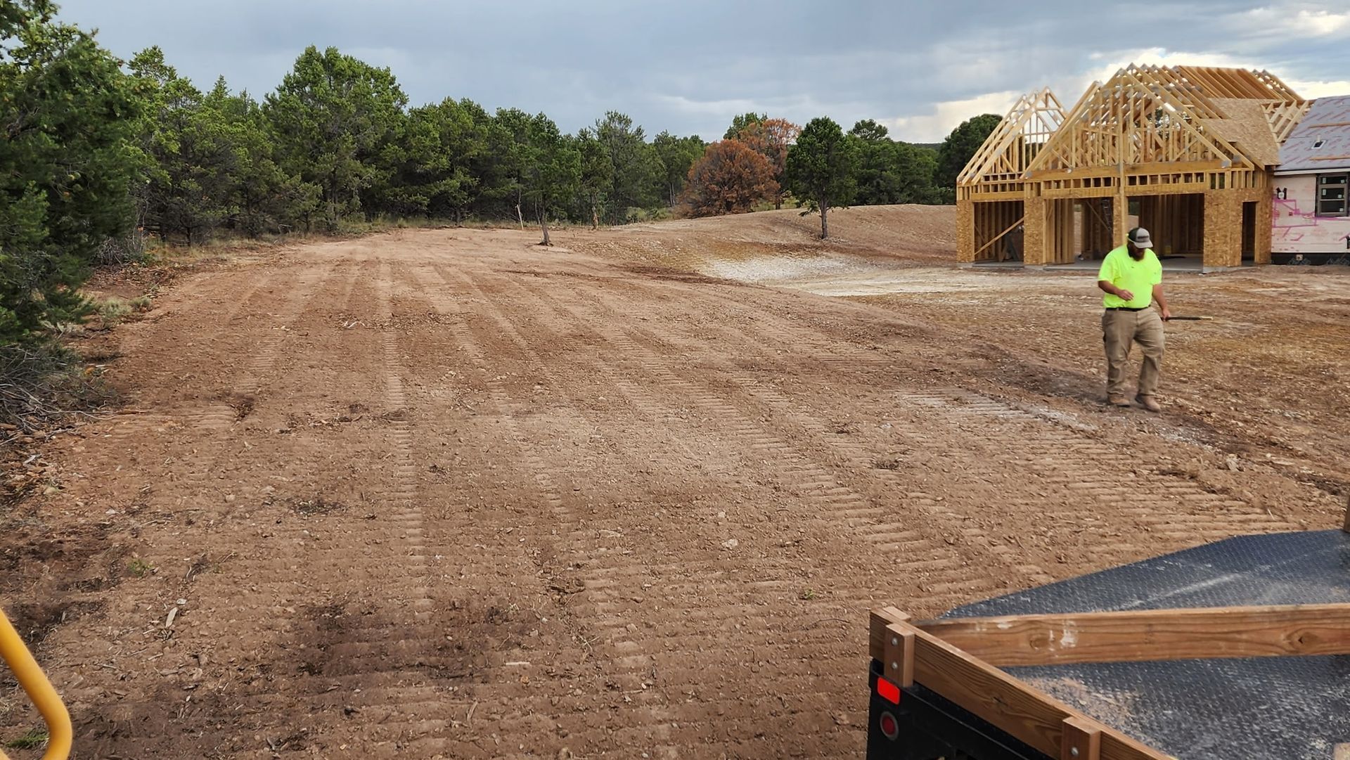Construction site with cleared dirt lot, trees, and a worker in a neon vest near a framed house under construction