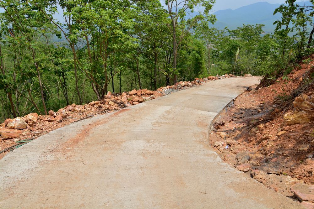 Concrete road winding through a wooded hillside with exposed red soil and distant mountains.