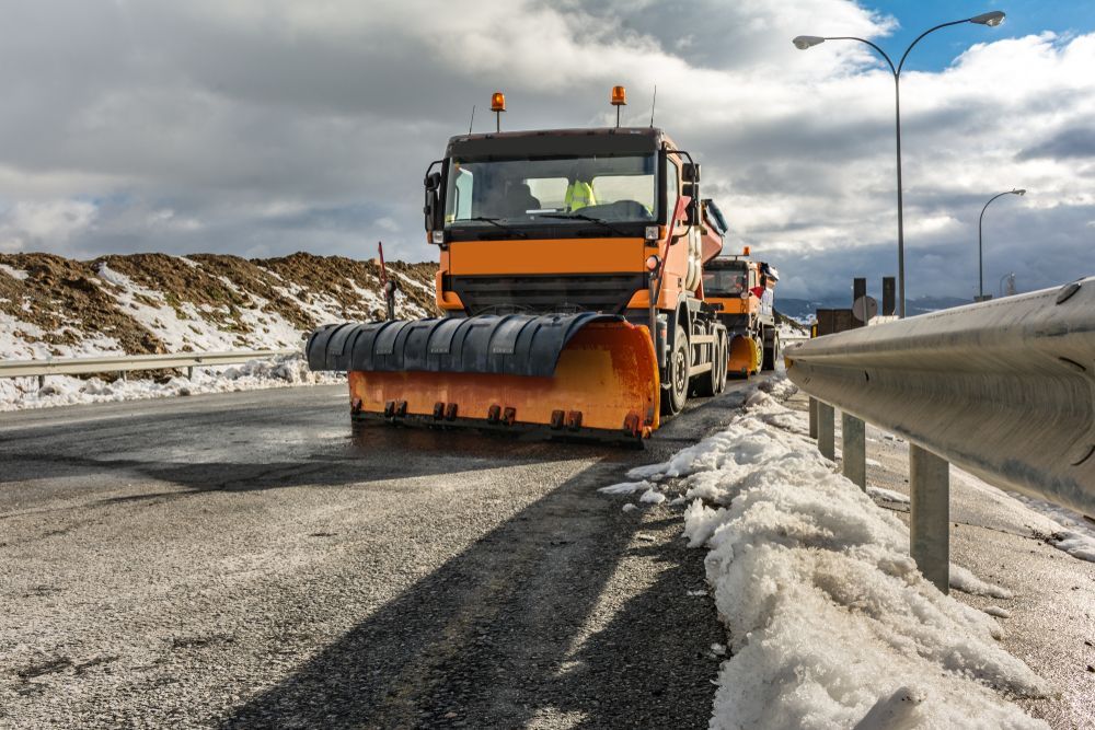 Orange snowplow clearing a snowy highway beside a concrete barrier under cloudy skies