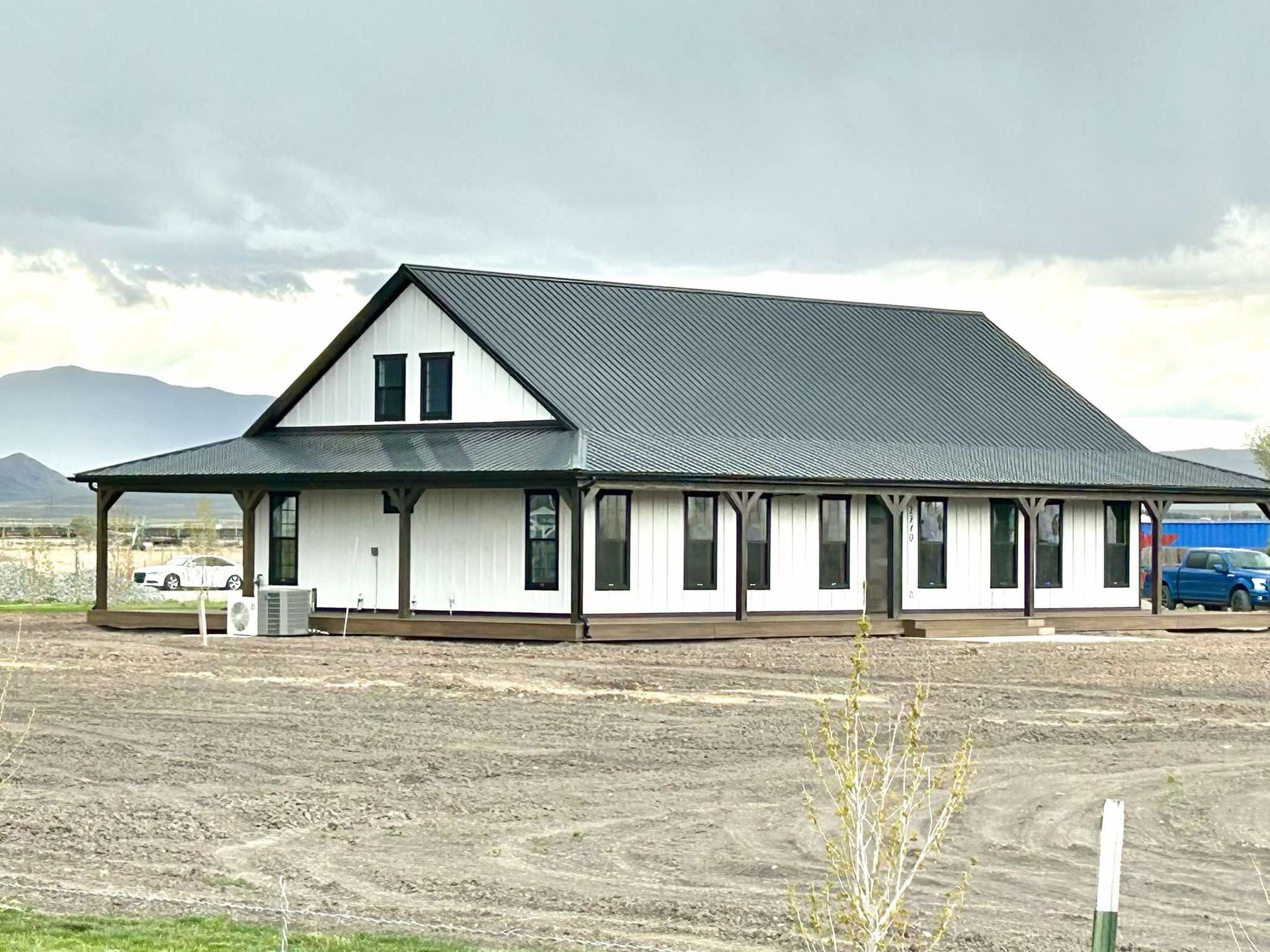 A large white barndominium with a black roof is sitting in the middle of a dirt field.