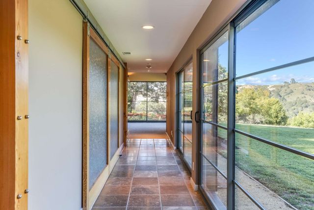 Modern hallway with terracotta tile, glass walls, and views of a green yard and hills.