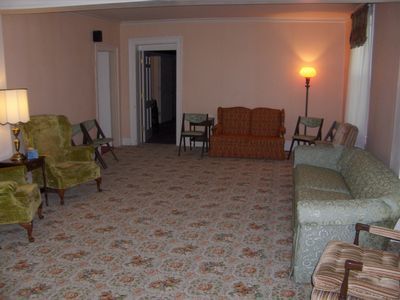 Empty living room with patterned carpet, sofas, armchairs, and warm lamps along the walls