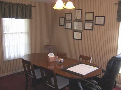 Dining room with wooden table and chairs, framed certificates on striped wall, and a window with curtains.