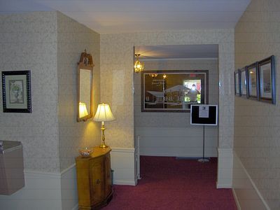 Hallway with beige walls, red carpet, lamp on a small cabinet, and framed pictures on the walls.