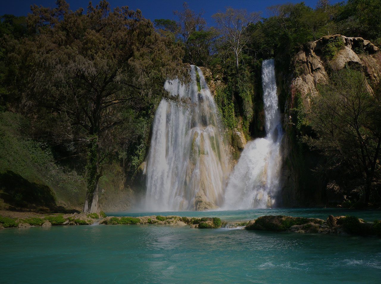 Una persona practica paddleboarding frente a una gran cascada en un entorno tropical exuberante.