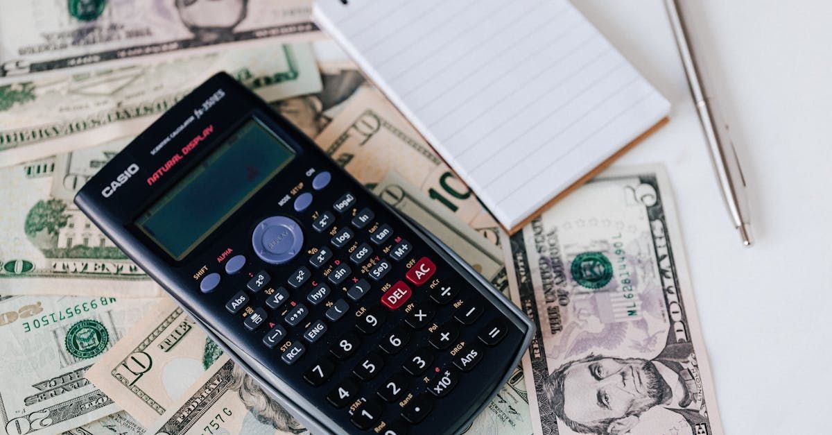 Calculator, notepad, and pen on a pile of US dollar bills, likely for financial calculations.