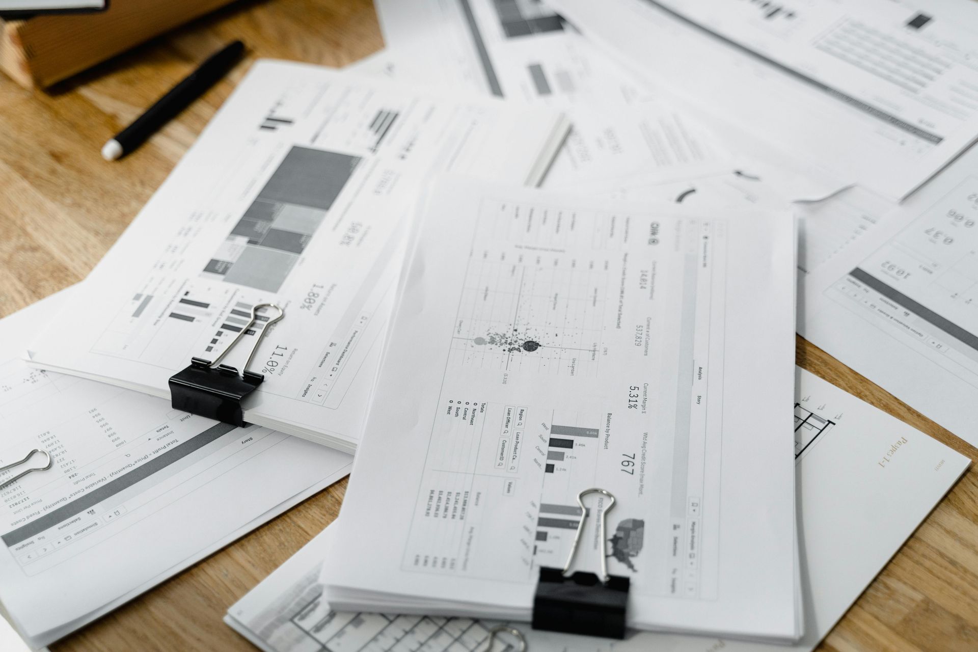 Papers with data and charts, held together with binder clips, scattered on a wooden surface with a pen.