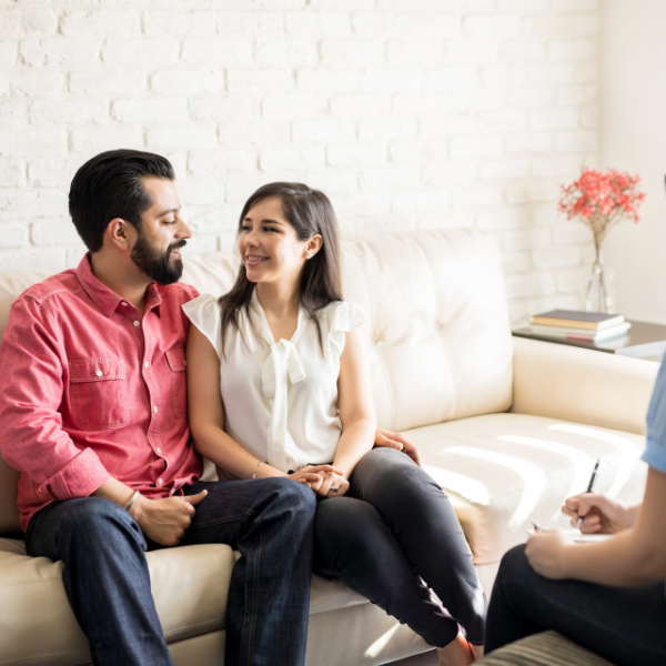 Two people sit on a beige couch, smiling and talking in a bright living room.