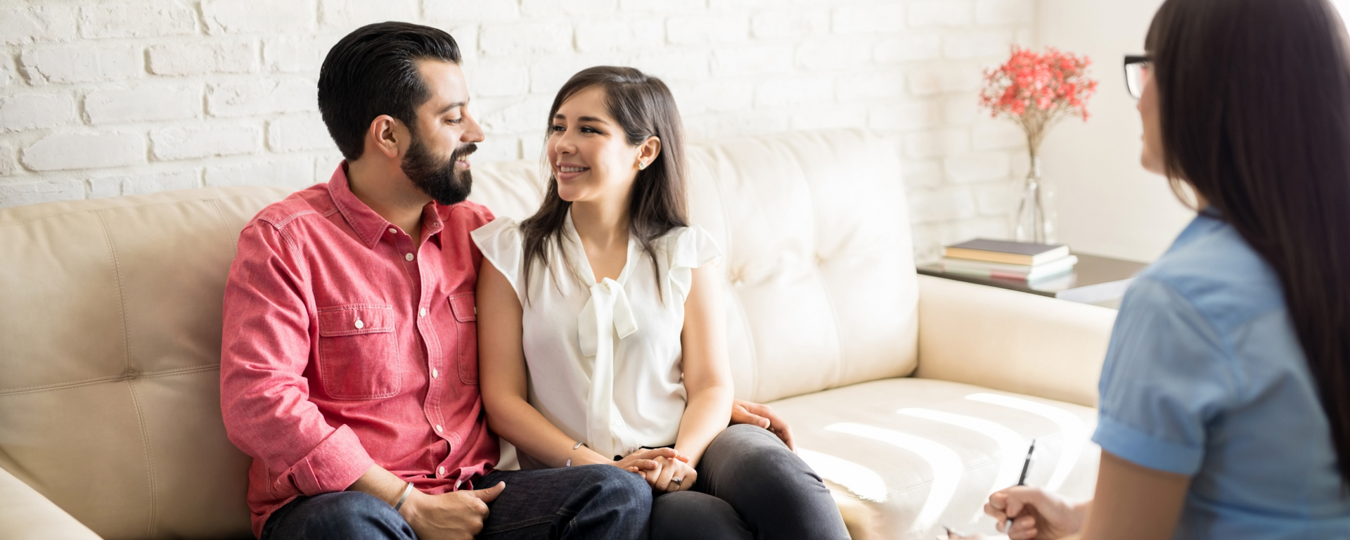 Two people sitting on a couch talking, with a third person nearby in a bright living room.