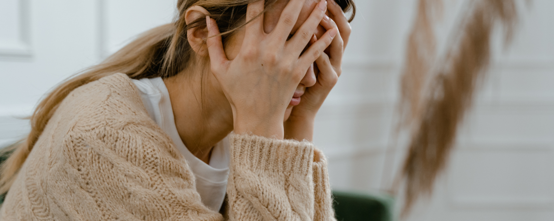 Person in a beige sweater covering their face, seated indoors in soft natural light