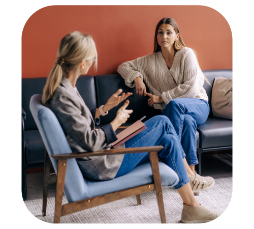 Two women talking in a cozy counseling office, seated on chairs and a sofa against a rust-colored wall.