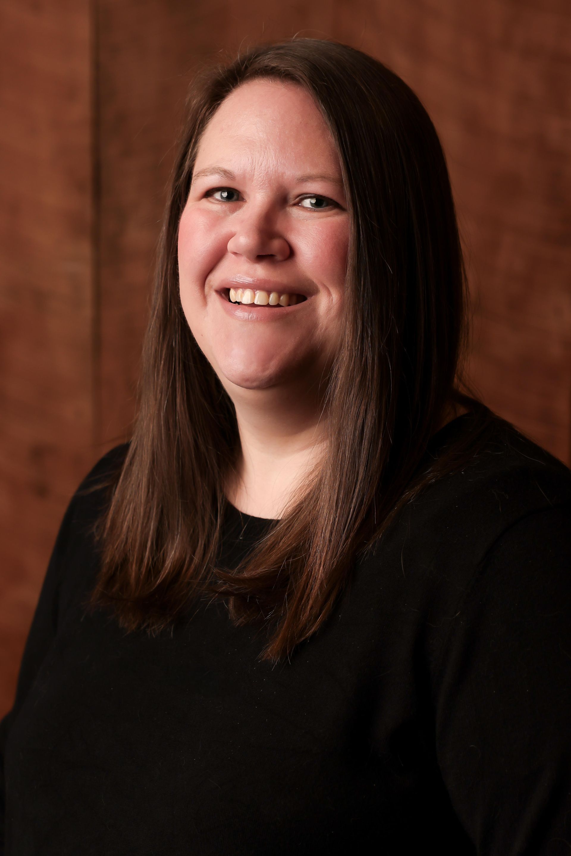 Portrait of a smiling person with long brown hair, wearing a black top against a warm brown backdrop