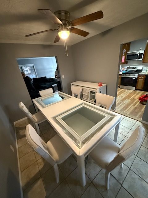 White dining table with glass inserts, four white chairs, gray walls, ceiling fan.
