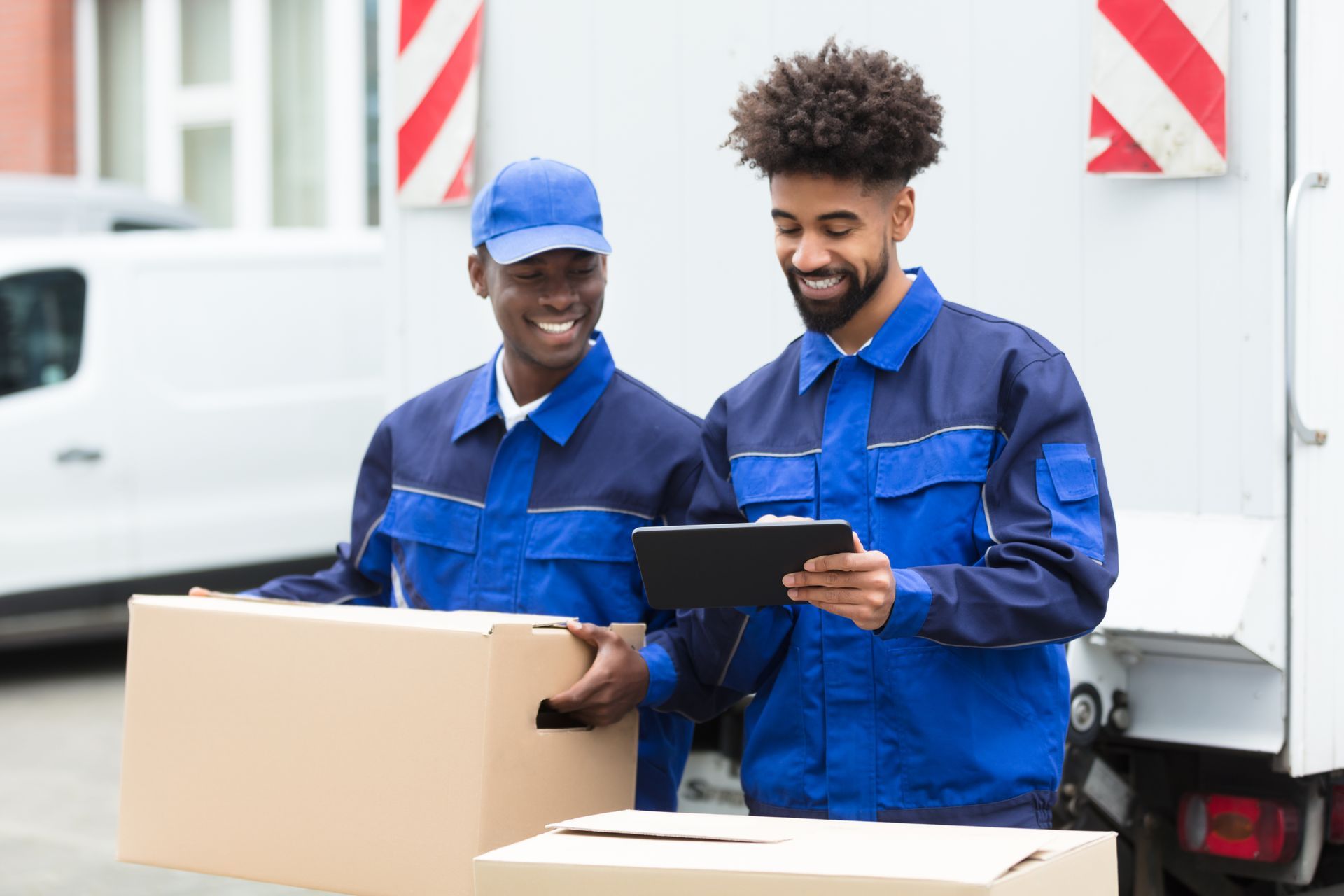 Two workers in blue uniforms, one holding a box, the other looking at a tablet near a truck.
